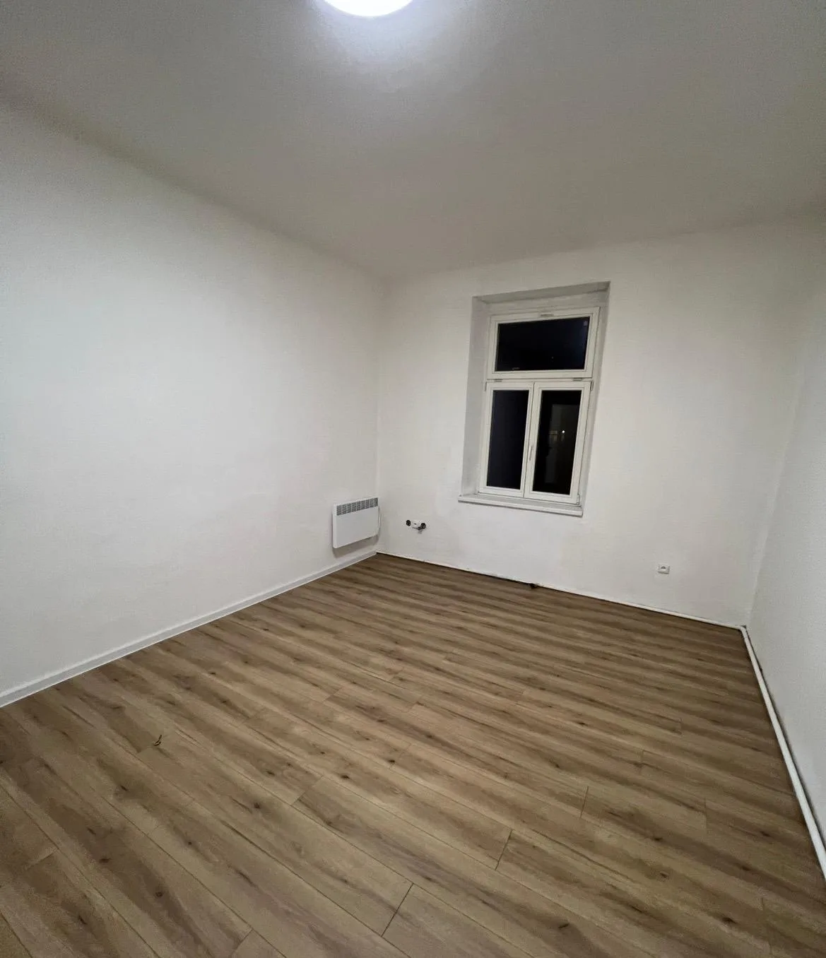 Vymalovaný pokoj v bílé barvě s tmavší dřevěnou podlahou, foceno večer.