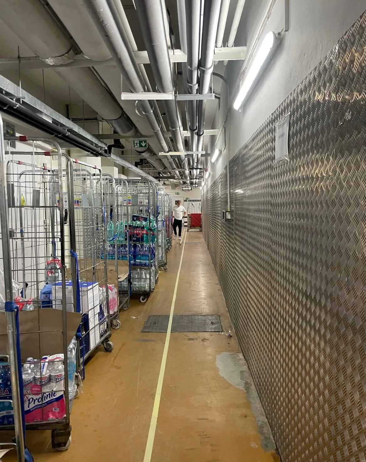 Servisní chodba komerčního objektu s kovovým obkladem a žlutým značením podlahy.
